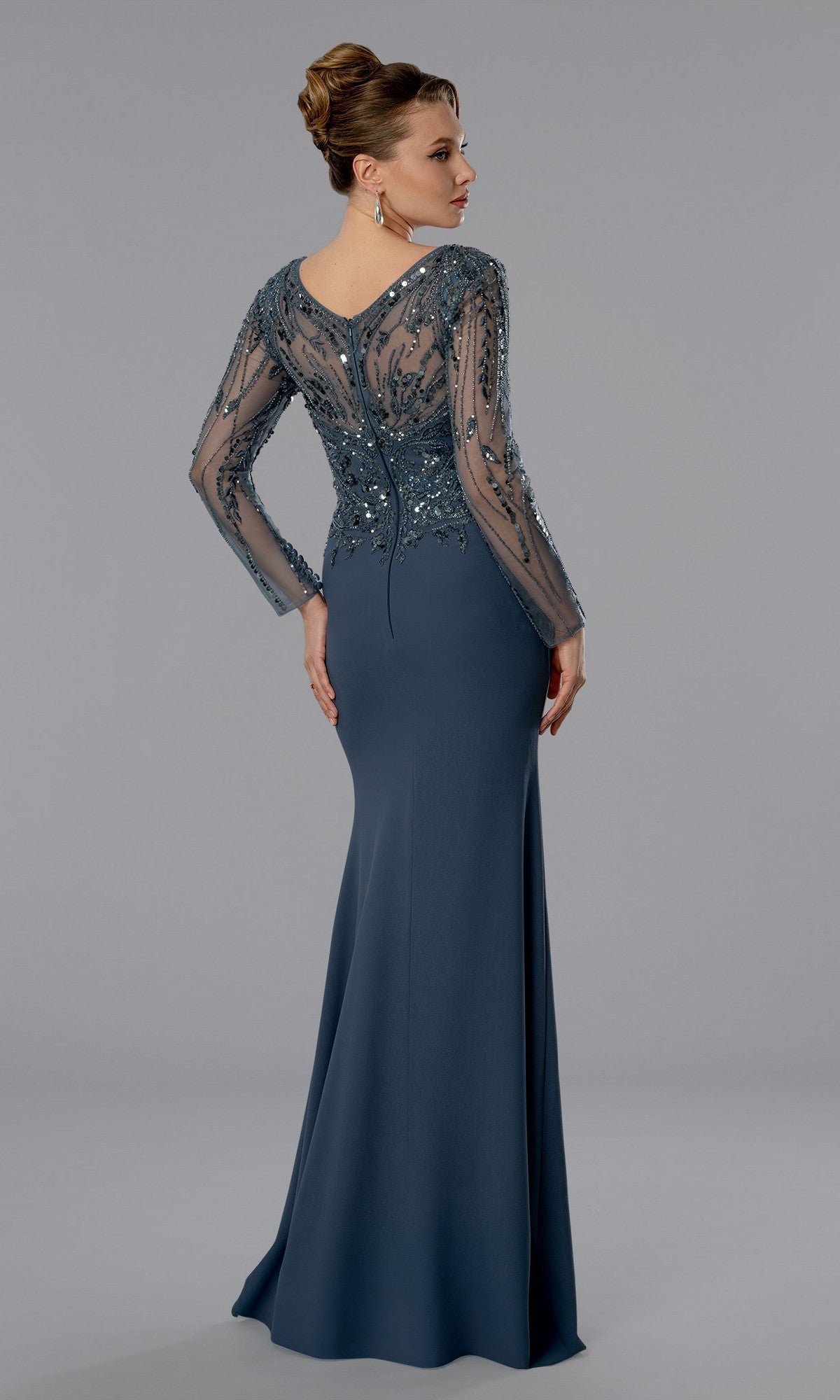 A woman wearing the Stella Couture 25371 long formal dress with sheer, beaded long sleeves and a fitted bodice stands facing away from the camera against a plain gray background.
